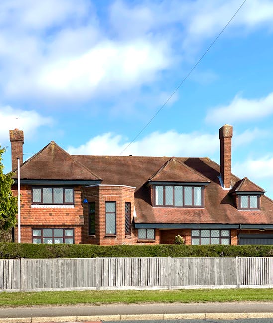 A large red brick house with a pitched roof and multiple chimneys, situated behind a wooden fence along a suburban street. The house features several rectangular windows, some with grid-patterned panes, and a central bay window on the ground floor. The property is surrounded by a well-maintained lawn and a hedge. The sky above is mostly blue with a few scattered clouds. This residential building exemplifies typical home architecture in Finchley, which Removal Van Finchley may assist with during house removals or relocation services, including furniture transport and packing. The photograph captures the exterior environment where moving trucks and boxes might be positioned during a home relocation process.