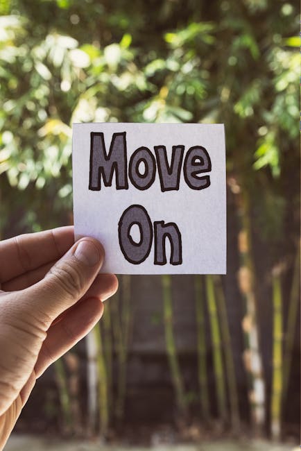 A person's hand holding a small white square paper with the words 'Move On' written in bold, black and purple lettering. The background features blurred green foliage and trees, suggesting an outdoor setting. The image conveys a message of transition or moving forward, which can be associated with home relocation or moving services. This visual is relevant for content about house removals, furniture transport, and packing and moving logistics, highlighting the concept of moving ahead during the home relocation process.