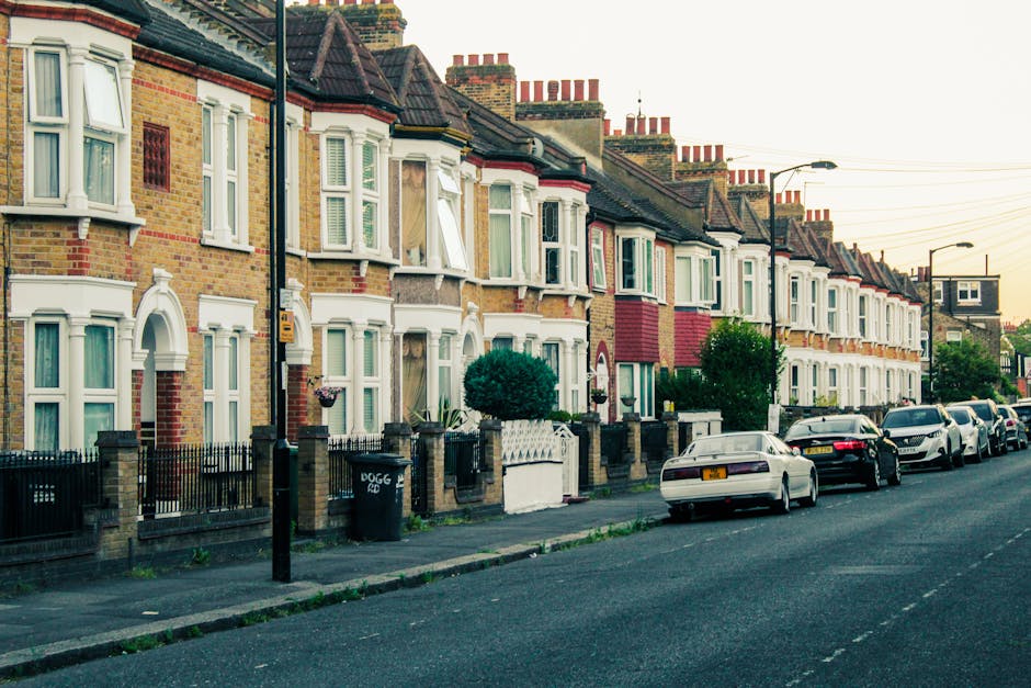 Image showing a row of terraced houses in a residential neighbourhood, with brick facades and bay windows, some with open window blinds. The houses have small front gardens enclosed by metal fences and brick walls; one garden features a neatly trimmed bush. Parked cars line the street in front of the houses, with a mix of different models and colours, including white, black, and silver vehicles. The street has a paved sidewalk, and there is a black wheelie bin labeled 'DOGG' positioned on the pavement. Street lamps are visible along the pavement, and overhead power lines run above the road. In the context of house removals, [COMPANY_NAME] may be involved in loading or transporting furniture and packing materials from these properties as part of a home relocation process, with the scene illustrating the typical environment where furniture transport and packing and moving activities occur.