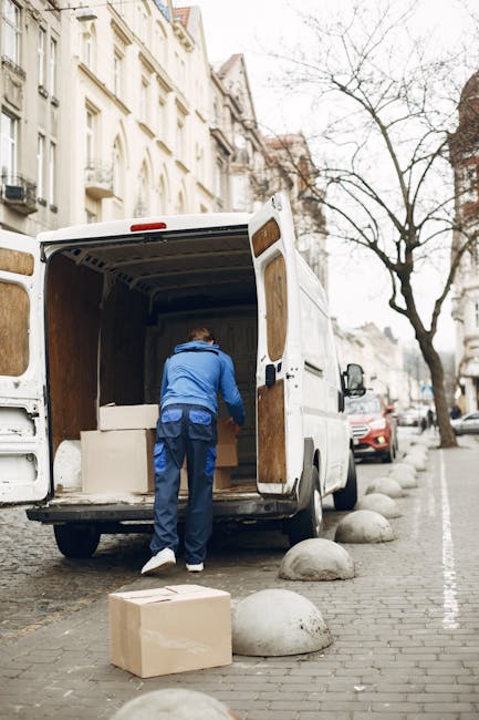 A man wearing a blue jacket and dark trousers is loading cardboard boxes and packaged furniture into the open cargo area of a white removal van parked on a city street. The van, associated with Removal Van Finchley, is positioned on a paved section of the pavement, with large rounded stone bollards separating it from the moving traffic and parked cars. Inside the van, visible wooden paneling and the interior space are used for transporting household belongings during a home relocation. The street backdrop features multi-storey buildings with classic architecture, leafless trees indicating a colder season, and overcast sky. The loading process includes the use of a trolley or dolly, and the scene captures the early stages of packing and moving, with the van part of a professional removals service.